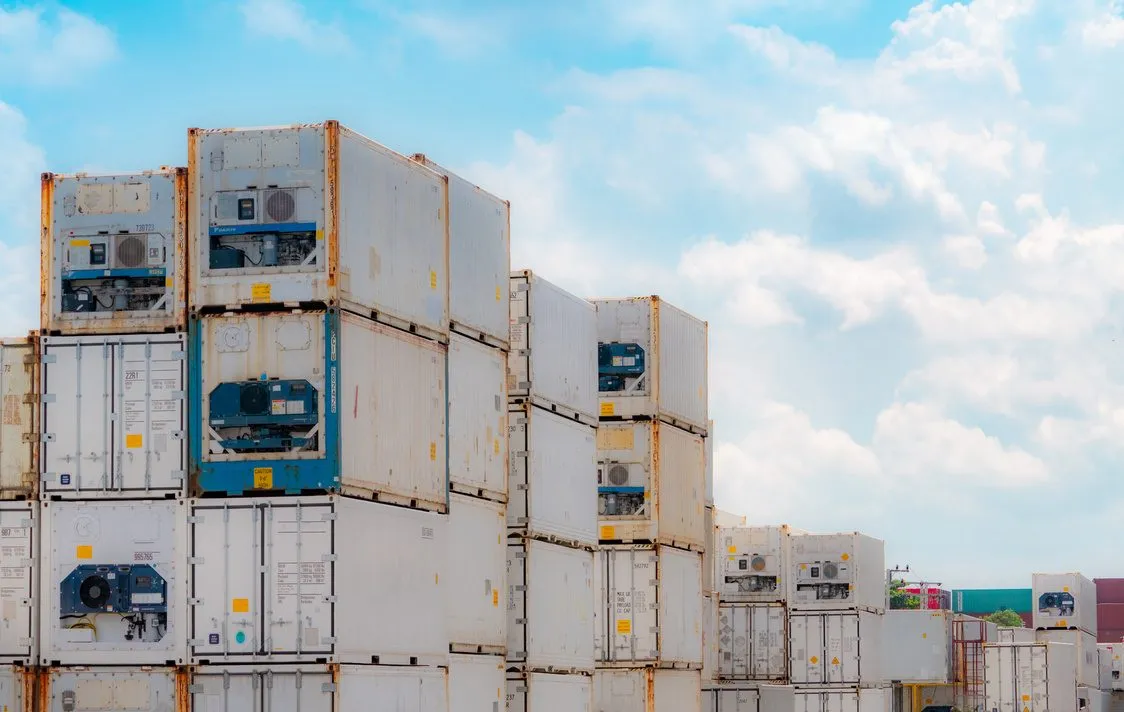Contenedores refrigerados blancos apilados en un patio, con los equipos de frío a la vista, bajo un cielo con nubes.